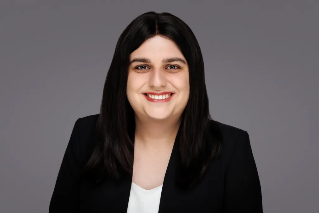 Smiling person in a black blazer, professional portrait against a gray background.