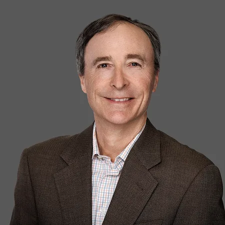 Smiling man in a brown suit jacket against a gray background.