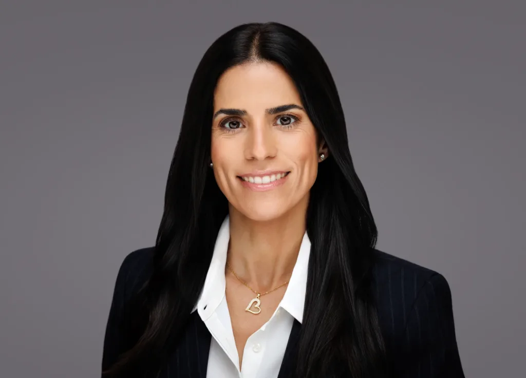 Smiling professional woman in a suit with long black hair and a heart necklace on a gray background.